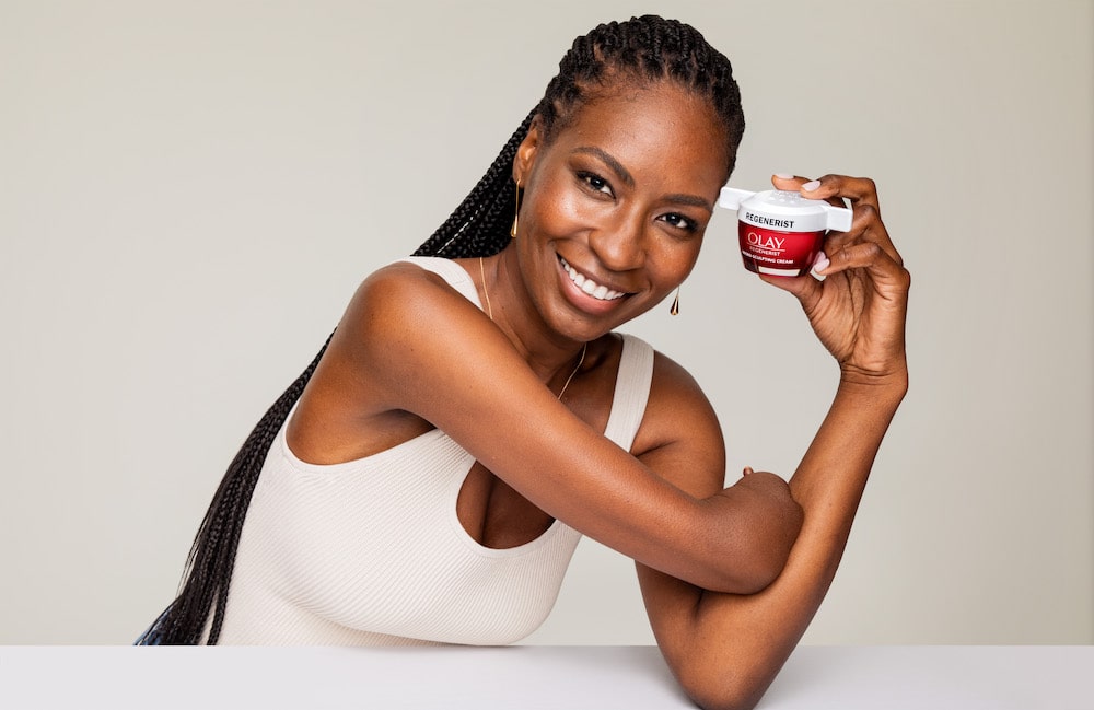 Olay Regenerist beauty product held by a Black woman in her left hand as she leans on her elbow. The lid is openable with one hand and the model with a limb difference smiles at the camera.